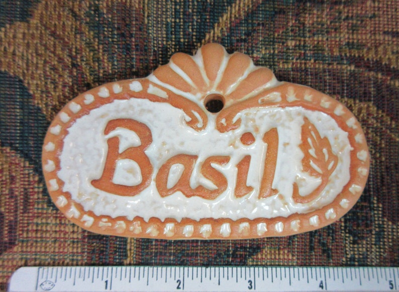 May include: A terracotta-coloured ceramic plant tag with a white glaze and the word "Basil" written in orange. The tag has a hole at the top for hanging.