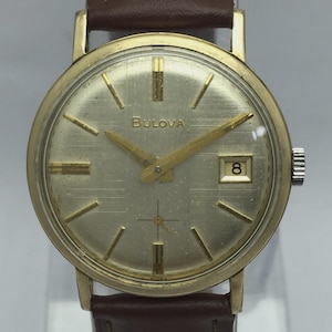 May include: Vintage Bulova wristwatch with a gold-tone case and a silver-colored dial. The watch features gold-tone hands and markers, a date window at the 3 o'clock position, and a brown leather strap. The watch face displays the Bulova brand name.