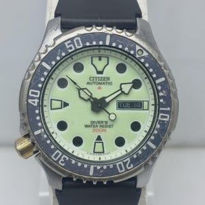 Puede incluir: Un reloj Citizen Automatic Diver's con una correa de caucho negra. La esfera es verde claro con marcadores y agujas negras. El bisel es azul oscuro con marcas blancas. El reloj muestra "TUE 14" y "DIVER'S WATER RESIST 200M".