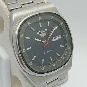 May include: A vintage Seiko 5 automatic wristwatch with a dark gray face. The watch has a stainless steel band and case. The face displays the day and date, with the day reading "FRI 26". The watch also has a 24-hour indicator.