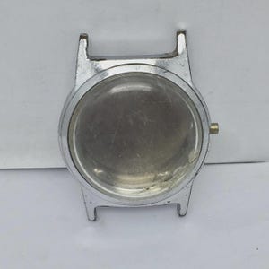 Może przedstawiać: Srebrna koperta zegarka z przezroczystą, okrągłą tarczą. Koperta posiada cztery uchwyty do mocowania paska i małą, złotą koronkę. Koperta zegarka jest pusta, bez elementów wewnętrznych.