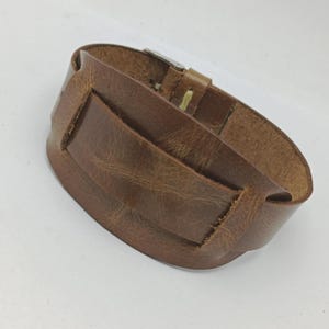 Peut inclure: Un large bracelet manchette en cuir marron avec une pièce d'accent rectangulaire. Le bracelet a une fermeture à boucle et est fait de cuir texturé et vieilli. Le bracelet manchette mesure environ 5 cm de large.