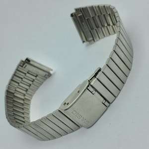 Puede incluir: Una correa de reloj extensible de color plateado con un cierre. La correa está hecha de eslabones metálicos rectangulares que están conectados entre sí. El cierre está marcado con la marca "CASIO". La correa está diseñada para un reloj.