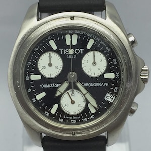 May include: A stainless steel Tissot chronograph watch with a black dial. The watch features white hands and markers, three sub-dials, and a date window. The watch is water-resistant to 100 meters or 330 feet. The text "TISSOT 1853" is visible.