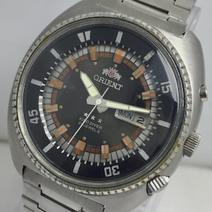 Puede incluir: Un reloj vintage Orient King Diver con una correa de acero inoxidable. La esfera negra tiene detalles en naranja y blanco, incluido el logotipo de Orient, la visualización del día y la fecha, y agujas luminosas. El bisel tiene marcadores de minutos.