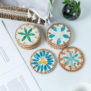 Op de afbeelding: Vier ronde onderzetters met bloemmotieven. Elke onderzetter heeft een geweven, lichtbruine rand. Het midden van elke onderzetter heeft een ander bloemmotief in groen, blauw en geel. De onderzetters liggen op een wit oppervlak.
