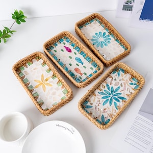 Op de afbeelding: Vijf rechthoekige geweven rotan trays met decoratieve interieurs. De trays hebben verschillende ontwerpen, waaronder bloemen- en vismotieven, met kleuren als blauw, groen en geel. Ideaal voor het serveren of presenteren van items.