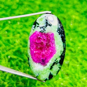 May include: An oval gemstone featuring a bright pink centre, encircled by a green and black speckled design. The gem is held by silver tweezers, set against a blurred green backdrop, highlighting its natural beauty and distinctive colour scheme.