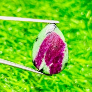 Peut inclure: Une pierre précieuse en forme de goutte d'eau tenue par une pince. La pierre présente un mélange de couleurs vives, avec un centre magenta foncé et une couche extérieure vert clair parsemée de noir. La pierre est sur fond vert vif.
