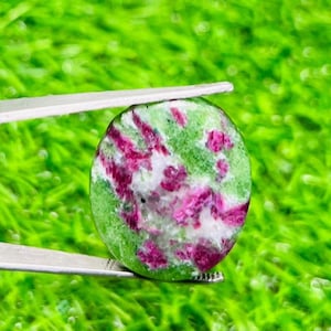 May include: A circular gemstone, held by tweezers, displaying a mix of green, white, and magenta hues. The polished stone is set against a bright green background, with a natural, organic appearance.