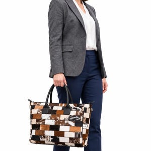 May include: A rectangular handbag featuring a patchwork design of brown, black, and white cowhide squares. The bag has two black leather handles and a top zipper closure, showcasing a multi-toned, textured finish with varied fur patterns.