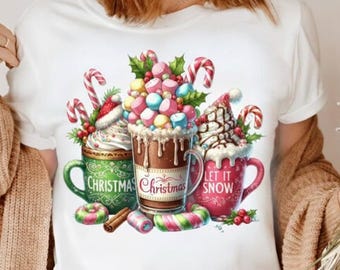 May include: White t-shirt featuring a festive graphic of three mugs of hot chocolate. Each mug is decorated with whipped cream, candy canes, and the words "Christmas" and "Let it Snow". Colorful marshmallows and sprinkles add to the holiday theme.