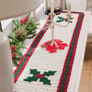 Puede incluir: Un camino de mesa de ganchillo color crema con un tema navideño festivo. El camino de mesa presenta un borde rojo y verde y diseños de hojas de acebo. Un candelabro plateado y flores de pascua rojas están sobre la mesa.
