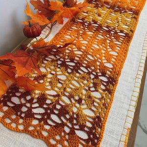May include: An orange and brown crocheted table runner with a leaf pattern. The runner is placed on a white linen surface and decorated with fall leaves and a small pumpkin. The color gradient transitions from dark brown to orange.