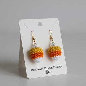 May include: A pair of handmade crochet earrings in the shape of candy corn. The earrings feature yellow, orange, and white sections, with gold-colored hooks and clear bead accents. The earrings are displayed on a white card with the text "Handmade Crochet Earrings".