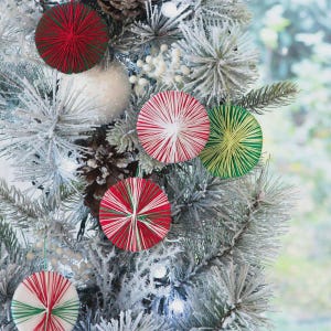 May include: Christmas tree decorated with handmade ornaments. The ornaments are round, made of red, white, and green thread, and arranged on a frosted evergreen tree. A white ball ornament and pine cones are also visible.