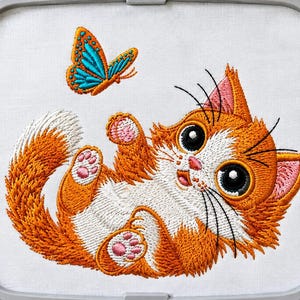 May include: An embroidery design featuring a fluffy orange and white kitten lying on its back with its paws raised. A bright blue and orange butterfly is positioned above the kitten. The entire design is contained within a gray embroidery hoop.
