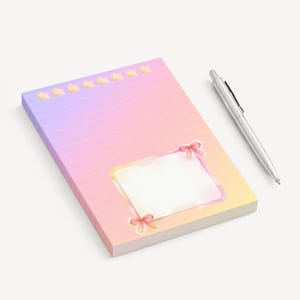 Peut inclure: Un bloc-notes aux couleurs pastel avec un dégradé de rose, de violet et de jaune, avec un cadre vide orné de nœuds roses et une rangée d'étoiles jaunes. Un stylo argenté est posé à côté du bloc-notes, prêt à écrire.