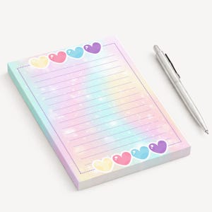Peut inclure: Un bloc-notes aux couleurs pastel avec un intérieur ligné et une bordure de cœurs colorés. Un stylo argenté repose à côté du bloc-notes. Le bloc-notes a une esthétique douce et rêveuse, idéale pour prendre des notes ou tenir un journal.