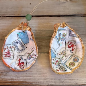 May include: Two decorative oyster shell ornaments. Each shell is painted with a gold rim and features colorful illustrations of playing cards and a pagoda. A green bead and twine are attached for hanging.