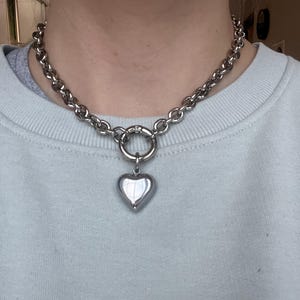 May include: A silver-toned chain necklace featuring thick, rounded links and a central circular clasp. A smooth, polished, puffed heart-shaped pendant hangs from the clasp, creating a bold, metallic look that rests against the collarbone area.
