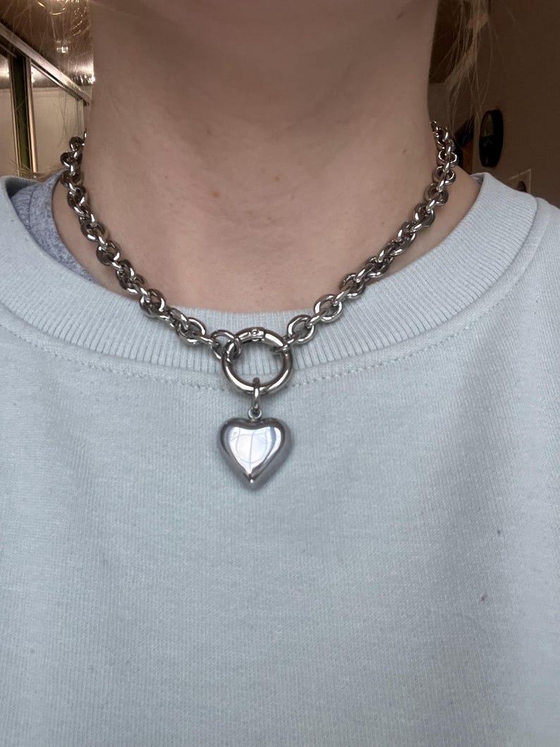May include: A silver-toned chain necklace featuring thick, rounded links and a central circular clasp. A smooth, polished, puffed heart-shaped pendant hangs from the clasp, creating a bold, metallic look that rests against the collarbone area.