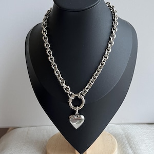May include: A silver-toned chain necklace featuring thick, rounded links and a circular clasp at the centre. A smooth, polished silver-toned heart-shaped pendant hangs from the circular clasp, creating a bold and reflective metallic appearance.