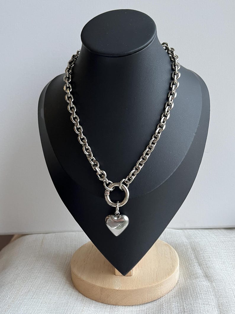 May include: A silver-toned chain necklace featuring thick, rounded links and a circular clasp at the centre. A smooth, polished silver-toned heart-shaped pendant hangs from the circular clasp, creating a bold and reflective metallic appearance.