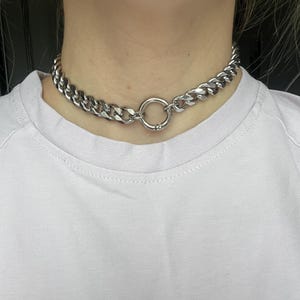 Op de afbeelding: Een zilverkleurige choker ketting. De ketting heeft een dik ontwerp met een curb chain met een grote cirkelvormige sluiting in het midden. De sieraden worden om de nek gedragen.
