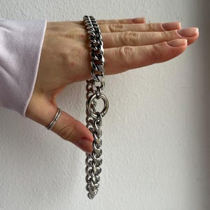 Op de afbeelding: Een zilverkleurige ketting met een dikke schakelontwerp. De ketting heeft een grote cirkelvormige ring in het midden. De sieraden worden gepresenteerd tegen een neutrale achtergrond.