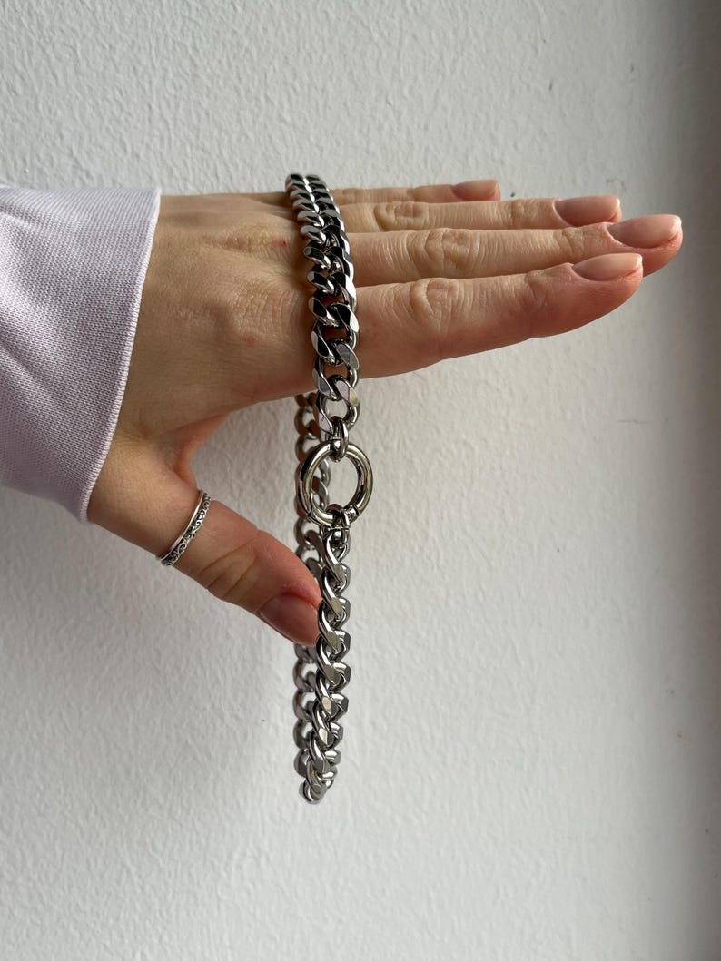 Op de afbeelding: Een zilverkleurige ketting met een dikke schakelontwerp. De ketting heeft een grote cirkelvormige ring in het midden. De sieraden worden gepresenteerd tegen een neutrale achtergrond.
