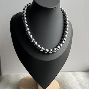 May include: A silver beaded necklace displayed on a black jewelry bust. The necklace is composed of numerous round, shiny beads. The bust is mounted on a wooden base, set against a neutral background.
