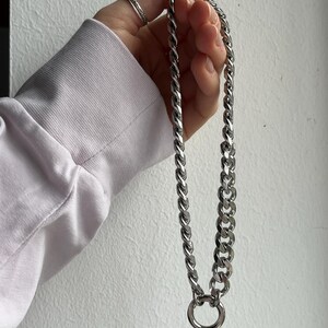 Op de afbeelding: Een zilverkleurige ketting met een grote cirkelvormige hanger. De ketting wordt vastgehouden door een persoon die een lichtpaars shirt met lange mouwen draagt. De schakels van de ketting zijn dik en verbonden. De persoon draagt ook een zilveren ring.