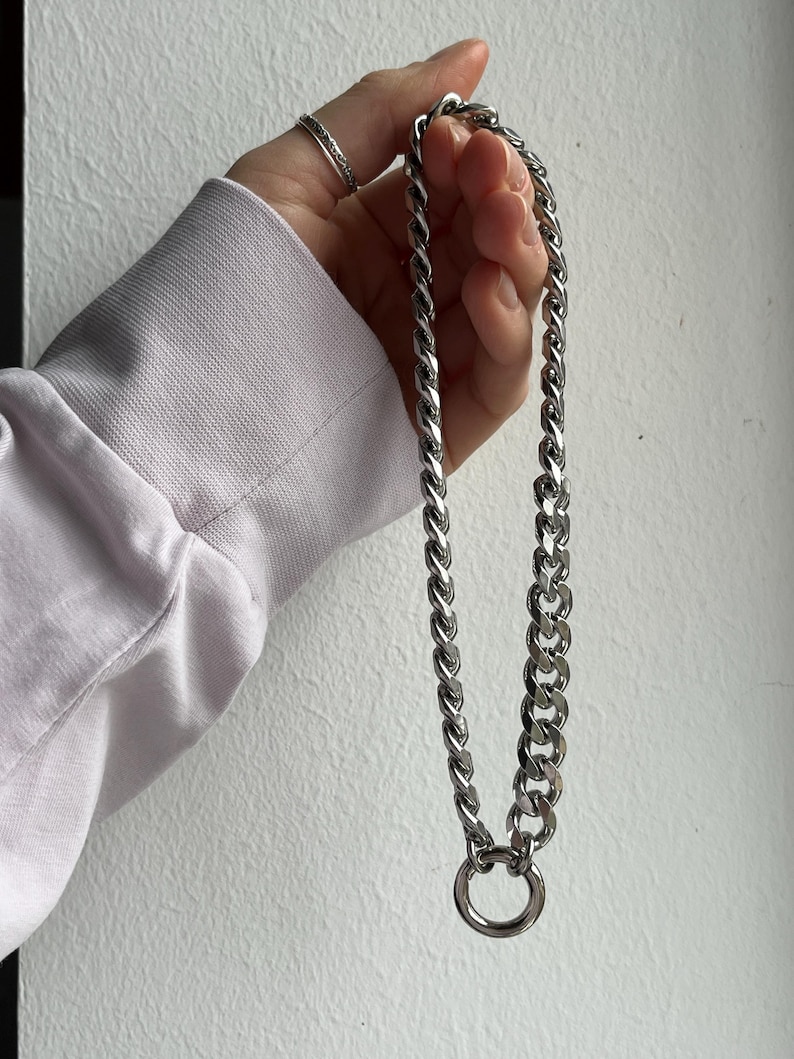 Op de afbeelding: Een zilverkleurige ketting met een grote cirkelvormige hanger. De ketting wordt vastgehouden door een persoon die een lichtpaars shirt met lange mouwen draagt. De schakels van de ketting zijn dik en verbonden. De persoon draagt ook een zilveren ring.