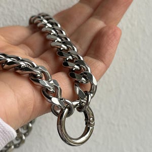 Op de afbeelding: Een zilverkleurige ketting met grote, in elkaar grijpende schakels. De ketting heeft een grote, ronde sluiting. De ketting wordt in een hand gehouden tegen een effen achtergrond.