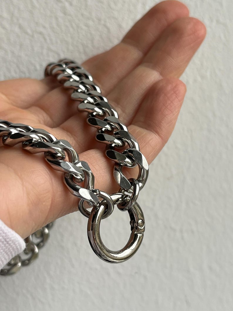 Op de afbeelding: Een zilverkleurige ketting met grote, in elkaar grijpende schakels. De ketting heeft een grote, ronde sluiting. De ketting wordt in een hand gehouden tegen een effen achtergrond.