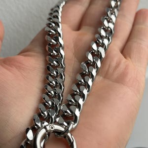 Op de afbeelding: Een zilverkleurige gourmette ketting met een grote sluiting en ring. De ketting is gemaakt van dikke, in elkaar grijpende schakels. De ketting wordt getoond tegen een neutrale achtergrond.