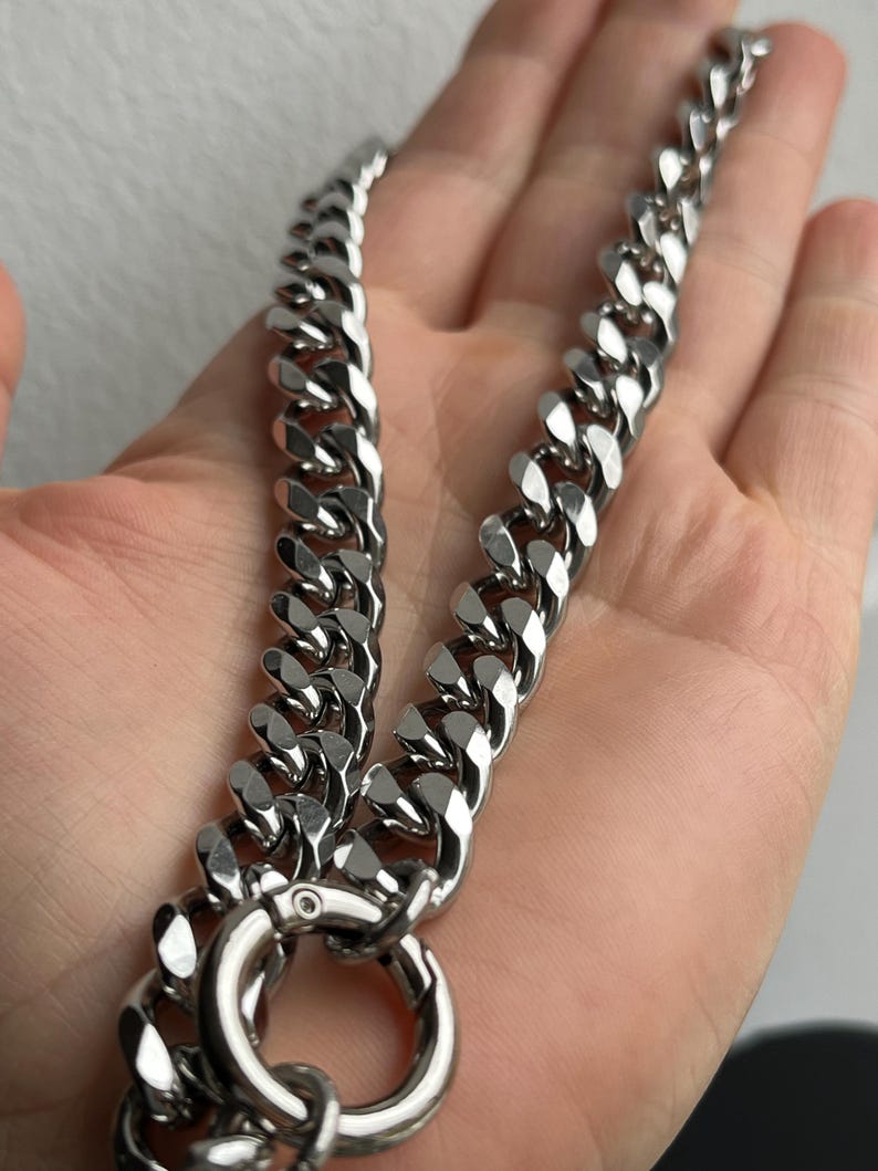 Op de afbeelding: Een zilverkleurige gourmette ketting met een grote sluiting en ring. De ketting is gemaakt van dikke, in elkaar grijpende schakels. De ketting wordt getoond tegen een neutrale achtergrond.