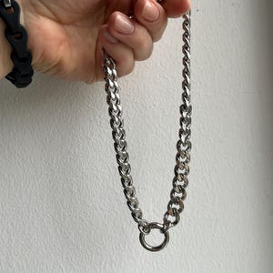 Op de afbeelding: Een zilverkleurige ketting met een grote, cirkelvormige sluiting. De ketting is gemaakt van dikke, in elkaar grijpende schakels. Op de achtergrond is een zwarte armband te zien.