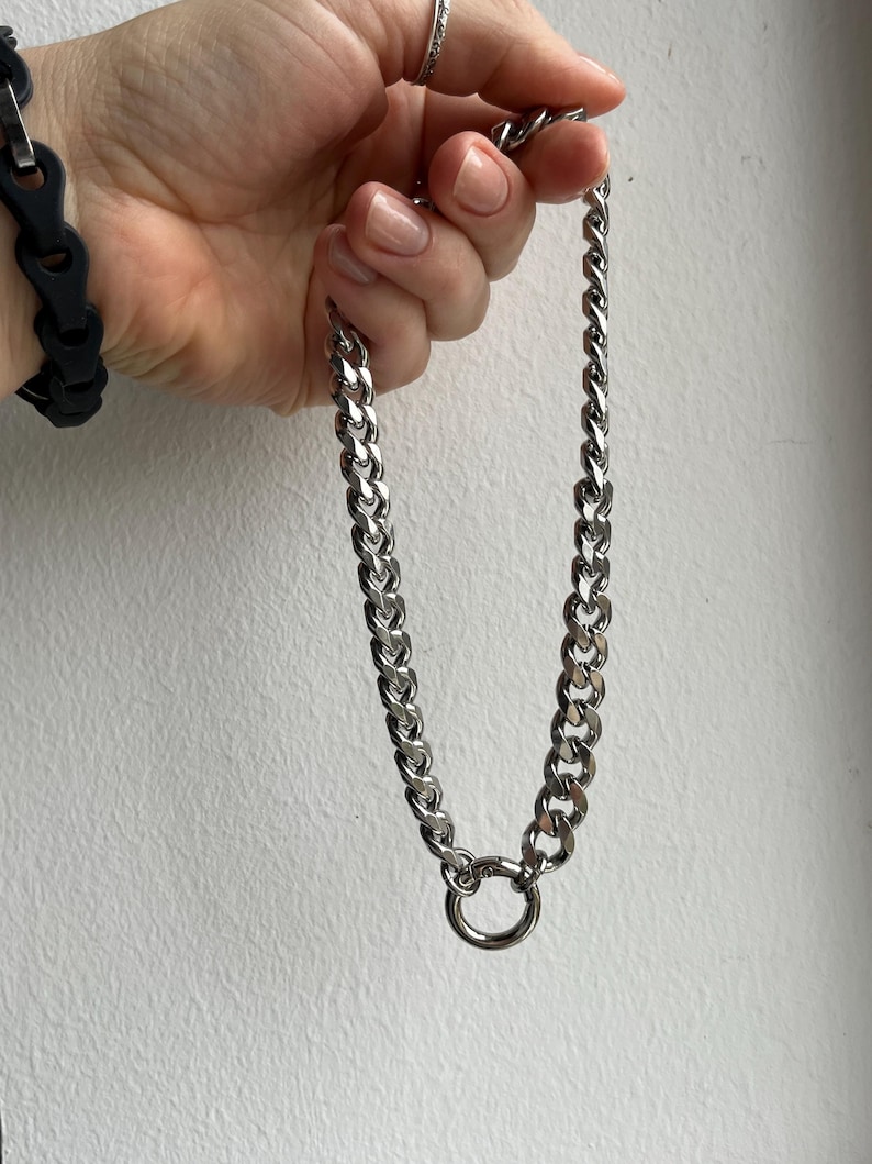 Op de afbeelding: Een zilverkleurige ketting met een grote, cirkelvormige sluiting. De ketting is gemaakt van dikke, in elkaar grijpende schakels. Op de achtergrond is een zwarte armband te zien.