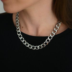Op de afbeelding: Een zilverkleurige gourmetketting met dikke, in elkaar grijpende ovale schakels met een gepolijste, reflecterende afwerking. De ketting rust op een zwart geribbeld shirt en toont een klassieke, stevige metalen constructie met een sluiting.