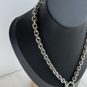 May include: A silver-toned chain necklace featuring thick, interlocking oval links. The necklace includes a circular clasp at the front, from which a smooth, polished silver-toned heart-shaped pendant hangs. The metal has a reflective finish.