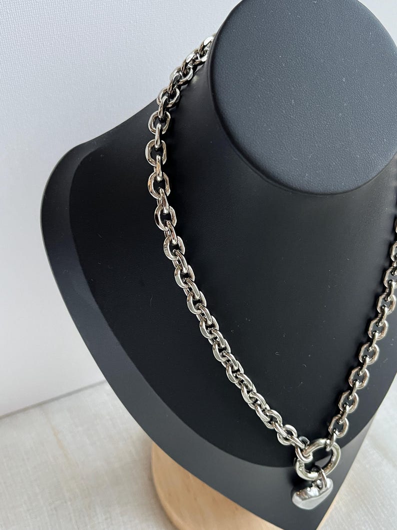 May include: A silver-toned chain necklace featuring thick, interlocking oval links. The necklace includes a circular clasp at the front, from which a smooth, polished silver-toned heart-shaped pendant hangs. The metal has a reflective finish.
