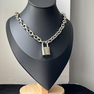 May include: A silver chain necklace with a padlock pendant, displayed on a black mannequin. The necklace has large, chunky links and a rectangular padlock. The mannequin is mounted on a wooden base.