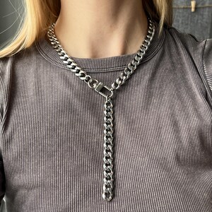 May include: A silver chain necklace with a chunky curb chain design. The necklace has a vertical chain drop with a square clasp. The jewellery is worn against a grey ribbed top.