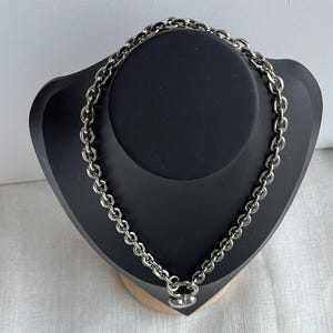 May include: A silver-toned metal chain necklace featuring thick, rounded interlocking links. The necklace is secured with a circular clasp at the bottom, which holds a small, smooth, heart-shaped pendant that hangs centrally from the chain.