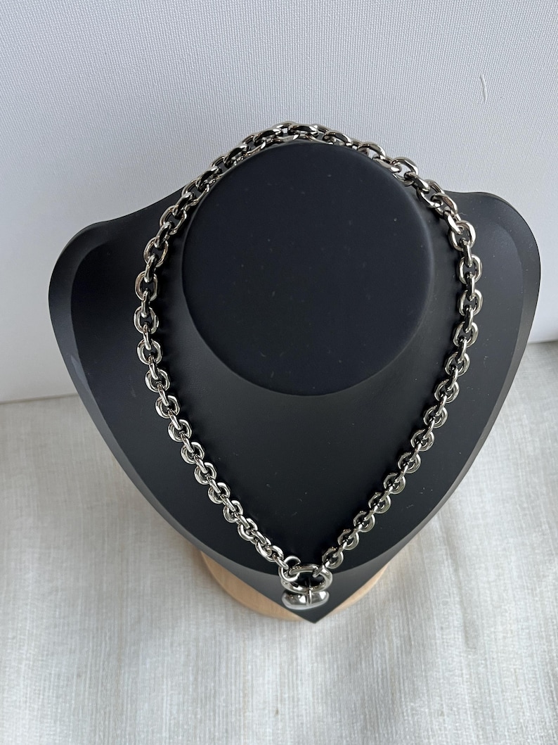 May include: A silver-toned metal chain necklace featuring thick, rounded interlocking links. The necklace is secured with a circular clasp at the bottom, which holds a small, smooth, heart-shaped pendant that hangs centrally from the chain.