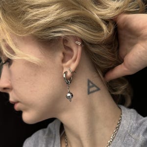 Puede incluir: Un pendiente de aro plateado con una bola plateada colgante y un pequeño pendiente de botón plateado son visibles en la oreja. También son visibles un brazalete de oreja plateado y un tatuaje de triángulo. La persona lleva una camisa gris y un collar de cadena plateada.