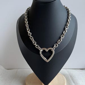 May include: Silver necklace with a heart-shaped clasp. The necklace has a chunky chain design and is displayed on a black mannequin. The heart clasp adds a romantic touch to the jewelry. The necklace is made of silver.