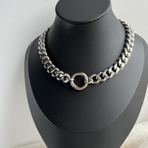 Op de afbeelding: Een zilverkleurige ketting met een grof geschakeld ontwerp. De ketting heeft een cirkelvormige sluiting in het midden en wordt gepresenteerd op een zwarte mannequin. Een opvallend sieraad.